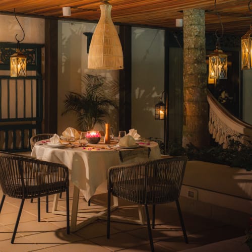 A cozy dinner setup with a round table, four chairs, and warm hanging lantern lights in a rustic indoor-outdoor setting.