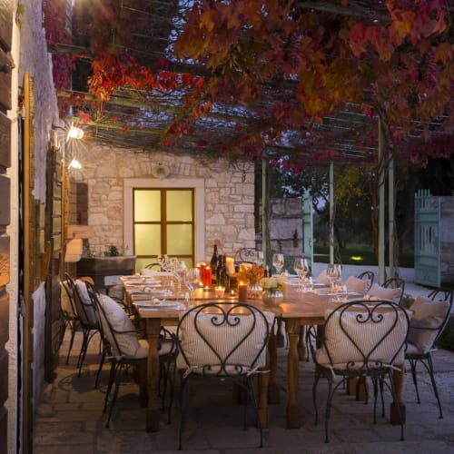 An outdoor dining setup on a terrace with a large wooden table, surrounded by cushioned wrought iron chairs, under a canopy of colorful autumn leaves, with candles and glassware on the table, in front of a stone building.