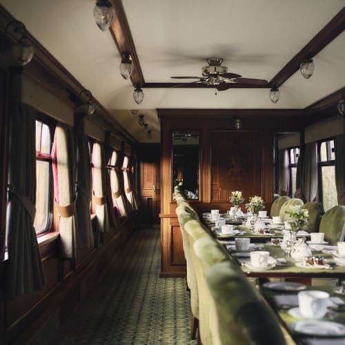 A vintage dining car interior with a long table set with white china and silverware, green upholstered chairs, and windows with dark curtains.