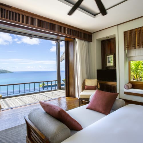 Ein luxuriöses Schlafzimmer mit großen Glastüren, die zu einem Balkon mit Blick auf den Ozean führen, mit Bett, Kissen, Sitzbereich und tropischer Landschaft.