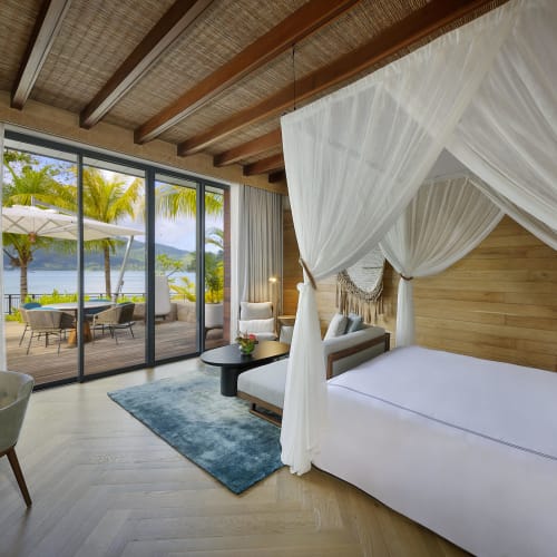 Luxuriöses Schlafzimmer mit großem Himmelbett, Holzbalkendecke und bodentiefen Glas Türen, die zu einer Terrasse mit Palmen und Blick auf das Meer führen.