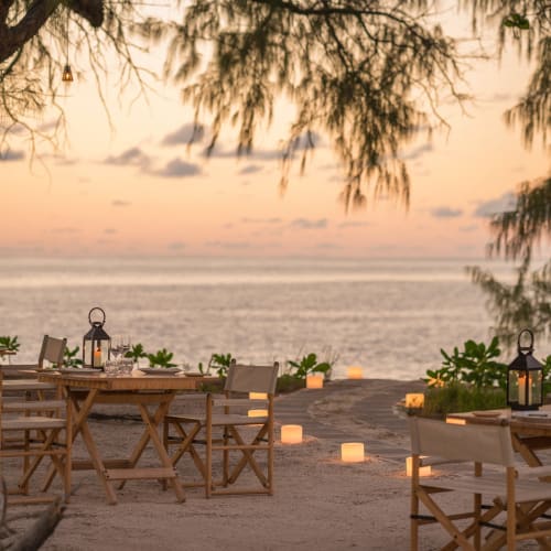 Außerhalb am Strand eingerichtete Essensplätze mit Holztischen und -stühlen, beleuchteten Laternen und Kerzen mit Blick auf einen ruhigen Ozean bei Sonnenuntergang.
