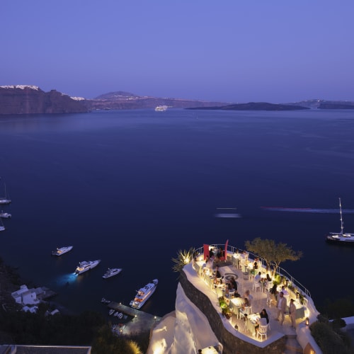 Evening view of a cliffside open-air restaurant with tables and guests, overlooking a calm sea with several anchored boats and a sailboat in the distance under a clear sky.