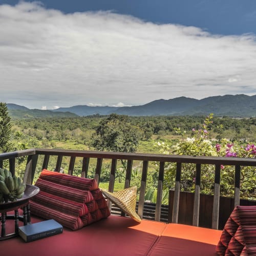 A cozy outdoor terrace with a cushioned seating area overlooking lush green hills and mountains in the distance, with blooming flowers and trees.