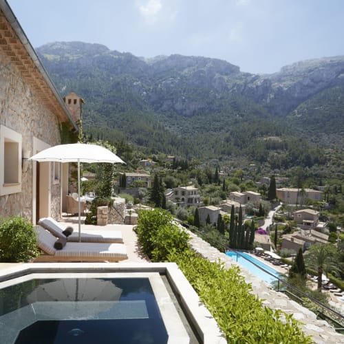 Eine Terrasse mit Liegestühlen und Sonnenschirm, die auf einen Hügel mit Häusern und üppigem Grün in Mallorca, Spanien, blickt.