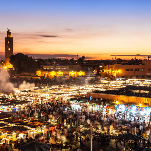 A vibrant night scene at Djemaa el Fna square in Marrakesh, bustling with market stalls, colorful lights, and crowds against the backdrop of a sunset sky.