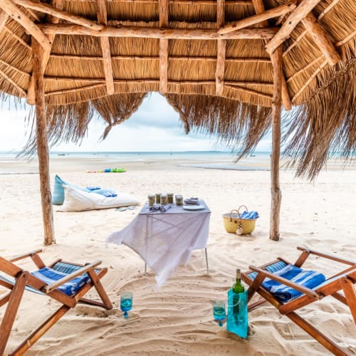 Ein schattiger Strandwagen mit zwei Liegestühlen, einem Tisch mit Getränken und Snacks, und das Meer im Hintergrund.