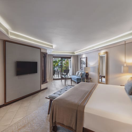 Modern hotel room with a large bed, a flat-screen TV mounted on the wall, seating area with sofa and coffee table, and a balcony with tropical plants visible outside.
