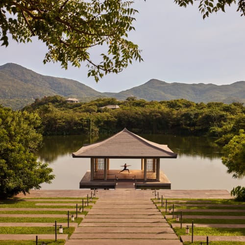 A serene lakeside pavilion with a person practicing yoga inside, surrounded by lush greenery and mountains.