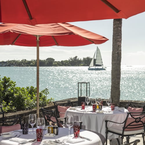 Outdoor restaurant tables with red umbrellas overlooking a sunlit sea and a sailboat near a distant shore with palm trees.