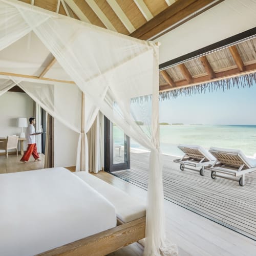 Wasser-Villa mit einem Baldachin-Bett, das auf das Meer blickt, mit zwei Liegestühlen auf dem Deck und einer Frau, die im Inneren der Villa geht.