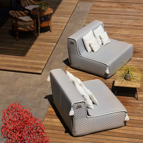 Outdoor-Terrasse mit modernen, gepolsterten Sesseln und kleinen Tischen auf Holzdielen, mit zwei gemusterten grauen Sesseln mit weißen Kissen und einer dekorativen roten Skulptur in der Nähe.