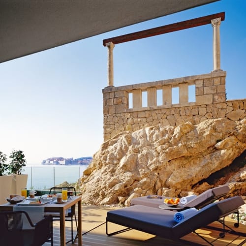 A sunlit terrace in a villa overlooking the sea with a table set for breakfast and a lounge chair.
