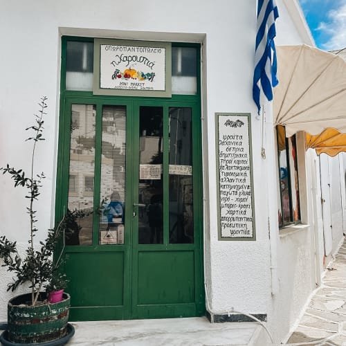 Entrance of a small marketplace with green doors in a white building, displaying Greek signage and a Greek flag hanging beside it under a sunny sky.