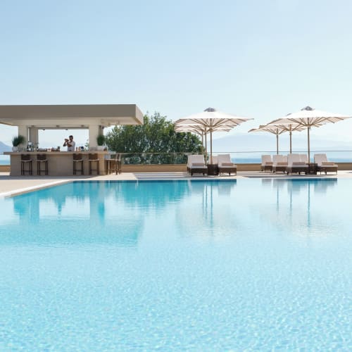 Ein ruhiger Außenpoolbereich mit Liegestühlen und großen Sonnenschirmen neben einer modernen Poolbar mit Blick auf das entfernte Meer unter klarem Himmel.