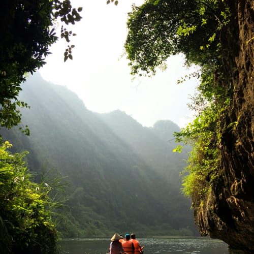 Zwei Personen in einem Ruderboot auf einem ruhigen Fluss, umgeben von steilen, bewaldeten Felsen und Sonnenstrahlen, die durch den Nebel in Ninh Binh scheinen.
