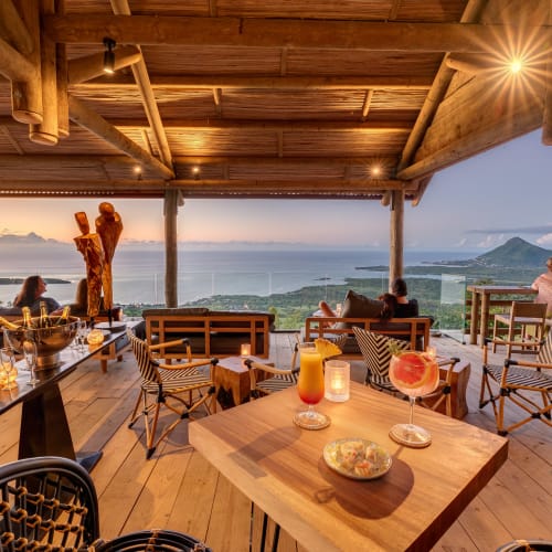 A cozy, open-air bar with wooden furniture and a view of the ocean and lush green hills at sunset.