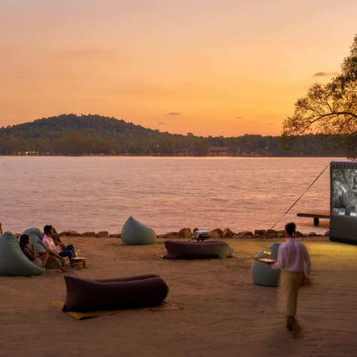 Outdoor cinema setup on a beach during sunset with people sitting on bean bags and a large screen showing a movie.
