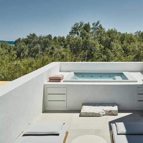 White outdoor terrace with a built-in jacuzzi surrounded by greenery under a clear sky.