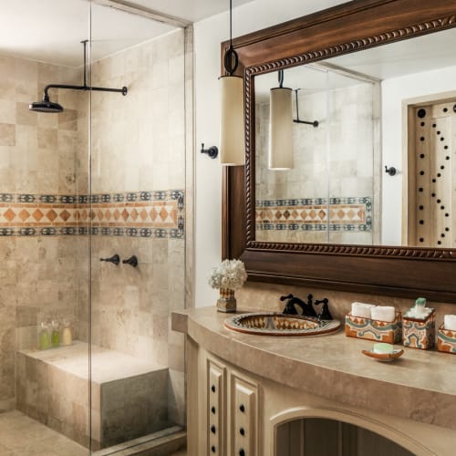 A luxurious bathroom with a large mirror above a marble countertop and a walk-in shower with beige tiles and decorative border.