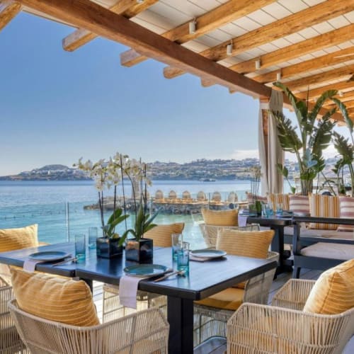 A seaside restaurant terrace with modern dining tables, colorful cushions, and lush green plants overlooking the water and distant islands.