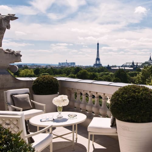 Ein eleganter Balkon mit Blick auf die Pariser Stadtlandschaft, einschließlich des Eiffelturms im Hintergrund. Der Balkon ist mit weißen Möbeln, Topfpflanzen und dekorativen Elementen ausgestattet.