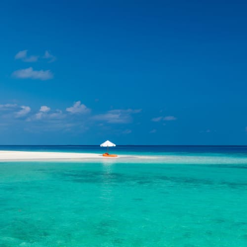 Eine ruhige tropische Strandlandschaft mit türkisfarbenem Wasser, weißem Sand, Palmen auf der linken Seite und einer kleinen Insel mit einem weißen Sonnenschirm im Hintergrund.