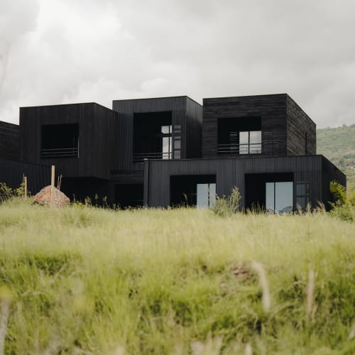 Modernes schwarzes Holzhaus mit kubischem Design hinter einem grasbewachsenen Feld unter bewölktem Himmel.
