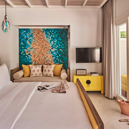 Modern bedroom with a large bed, a yellow accented bench against a blue and gold mosaic wall, a mounted flat screen TV, and sliding glass doors leading to an outdoor seating area with yellow couches.