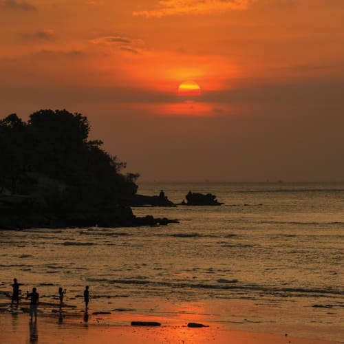 Sonnenuntergang über dem Meer mit Silhouetten von Bäumen und Menschen am Strand in Jimbaran, Bali.