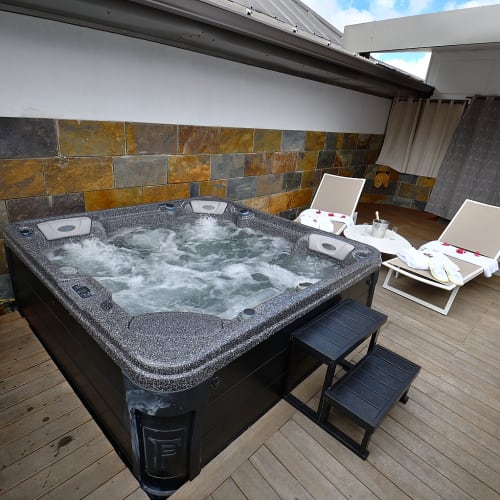 Außen-Whirlpool mit sprudelndem Wasser auf einem Holzdeck, begleitet von zwei weißen Liegestühlen mit zusammengerollten Handtüchern und Rosenblättern, umgeben von Pflanzen und einer Steinwand unter einem überdachten Bereich.