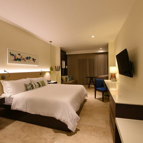 A hotel room with a large bed, wall-mounted TV, and a workspace near a window with curtains.