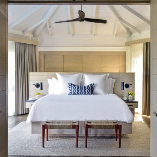 Ein luxuriöses Schlafzimmer mit großem Bett, weißer Bettwäsche und Dekokissen, in einem hellen, luftigen Raum mit hohem Decken und Ventilator, mit Blick auf das Wasser.