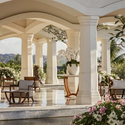 Offener Lobby-Lounge-Bereich mit weißen Säulen, Holzstühlen, Tischen und Topfpflanzen unter einem beigefarbenen gewölbten Dach, mit Blick auf ein Wasserbecken und blühende Sträucher im Garten.
