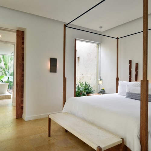 Luxurious overwater suite in Rosewood Mayakoba, with a large bed, white linens, and a sitting area visible through the doorway in the background.