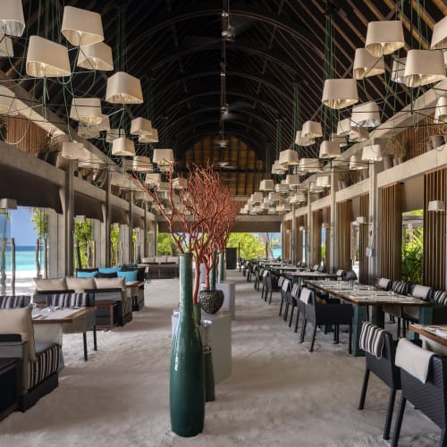 Freiluftrestaurant mit hohem gewölbtem Dach, modernen Hängeleuchten und Sitzgelegenheiten mit Blick auf einen Strand und das türkisfarbene Meer durch große Öffnungen.