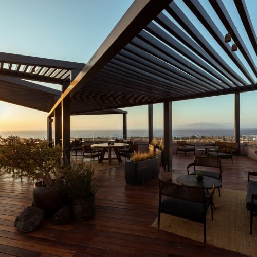 Große Dachterrasse mit modernen Pergolen, Lounge-Stühlen, Tischen und Topfpflanzen bei Sonnenuntergang mit Blick auf eine Küstenstadt.