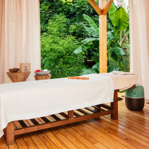 A massage table draped with a white sheet inside a serene room with large window overlooking lush greenery.