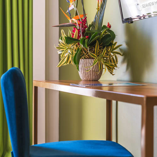 Eleganter blau gepolsterter Stuhl vor einem Holzschreibtisch mit einem lebhaften tropischen Blumenarrangement in einer neutralen Vase vor einer hellgrünen Wand mit einem teilweise sichtbaren Schild und grünen Vorhängen im Hintergrund.