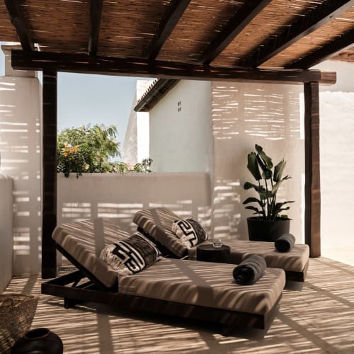 Eine schattige Dachterrasse mit zwei modernen Liegestühlen, jeweils mit gemusterten Kissen und zusammengerollten Handtüchern, unter einem Holzpergola mit einfallendem Licht.