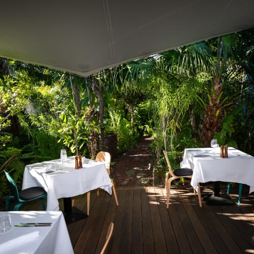 Außenrestaurant mit Tischen, die mit weißen Tischdecken bedeckt sind, umgeben von üppigen tropischen Pflanzen und beschattet durch ein Überdach.