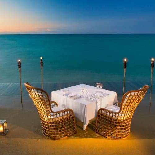 Ein romantisches Abendessen mit einem Tisch für vier Personen am Strand bei Sonnenuntergang, mit Blick auf das Meer, umgeben von Tiki-Fackeln und Laternen für stimmungsvolles Licht.