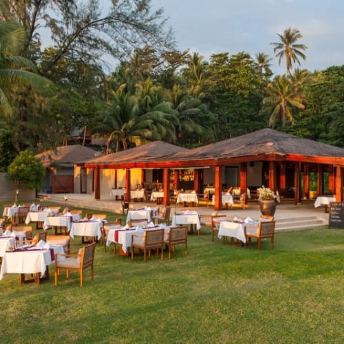 Outdoor dinner setting with tables and chairs on a grassy area in front of a tropical restaurant during sunset.