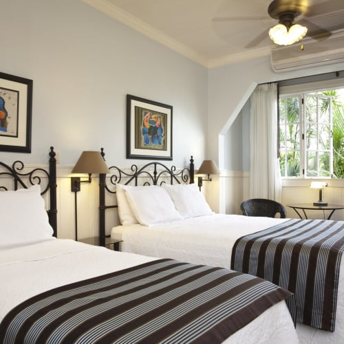 Zwei Einzelbetten in einem hellen Hotelzimmer mit weißen Bettdecken und gestreiftem Bettläufer, dekoriert mit gerahmtem Kunstwerk und einem großen Fenster mit Blick auf Pflanzen.