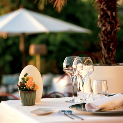 Tisch im Freien in einem Restaurant bei Tageslicht mit weißer Tischdecke, kleinem Blumentopf mit gelben Rosen, Weingläsern und einem weißen Sonnenschirm im unscharfen Hintergrund.