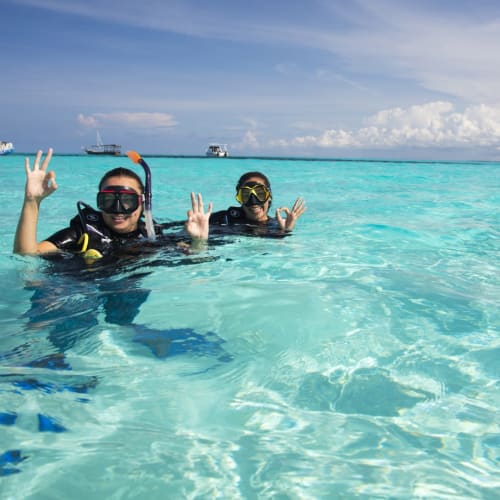 Zwei Personen mit Schnorchelausrüstung und Masken schwimmen im kristallklaren tropischen Wasser mit Booten im Hintergrund.