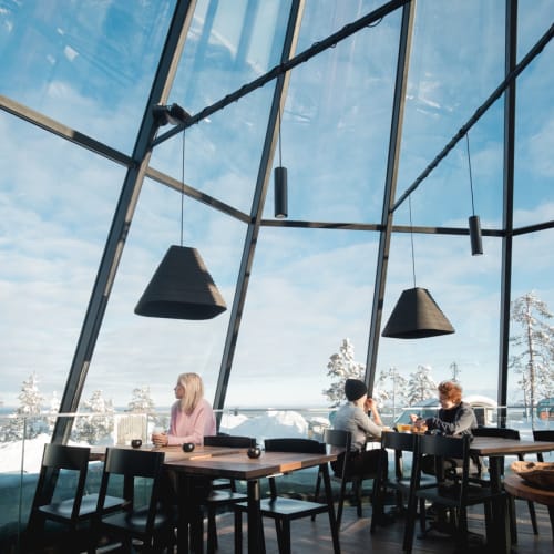 Drei Personen sitzen an Holztischen in einem Glas-Iglu-Restaurant mit großen schwarzen Hängelampen und einer verschneiten Landschaft draußen.