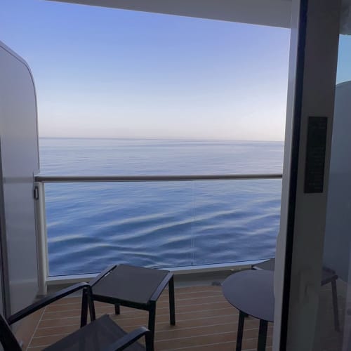 Kleiner Balkon auf einem Schiff oder Kreuzfahrtschiff mit zwei schwarzen Stühlen, einem runden Tisch und einer Glasgeländeransicht auf ruhiges blaues Meer unter klarem Himmel.