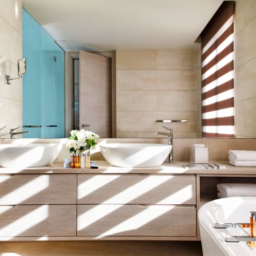 Helles Badezimmer mit Doppelwaschbecken, Badewanne und natürlichem Sonnenlicht, das durch gestreifte Fensterjalousien scheint.