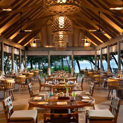 Elegantes Restaurant mit Holzmöbeln und reetgedecktem Dach, mit Blick auf einen tropischen Strand und den Ozean.